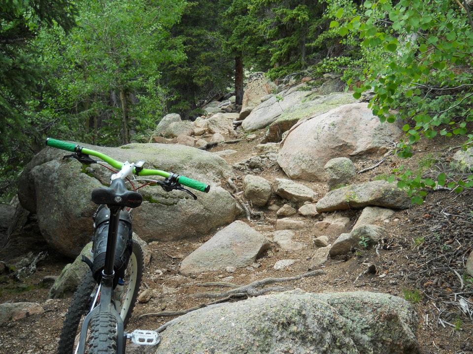 A mountain bike resting on rocky terrain, with a narrow path winding through large boulders and surrounded by lush greenery in a forested area. Barr Trail / Pikes Peak mountain bike trail.