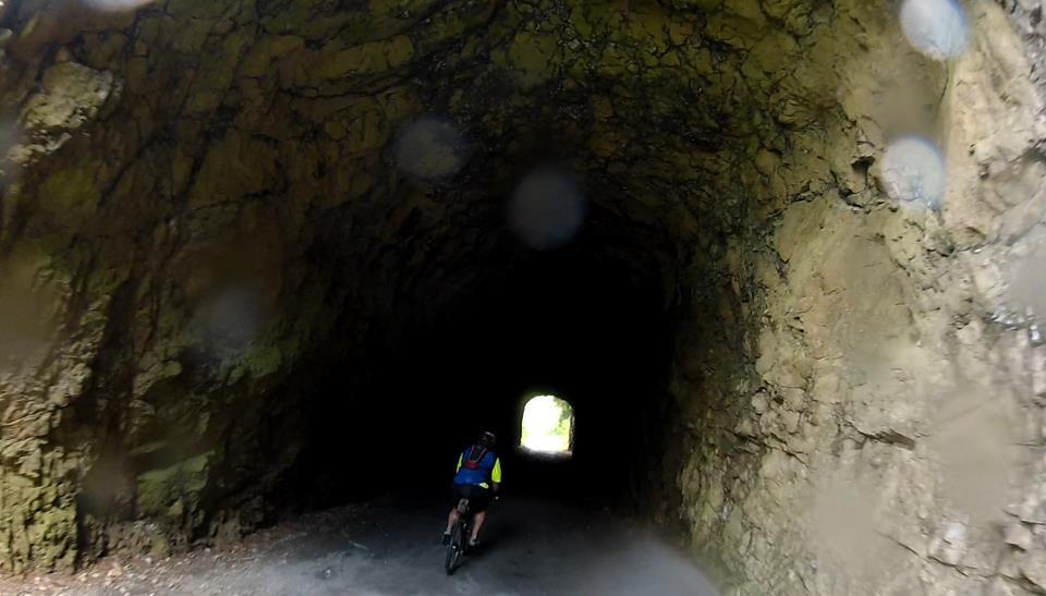 A cyclist riding through a dark tunnel with rocky walls, leading to a bright exit in the distance. The image has a slightly blurred appearance due to water droplets on the lens. New River Trail, Galax To Fries Junction mountain bike trail.