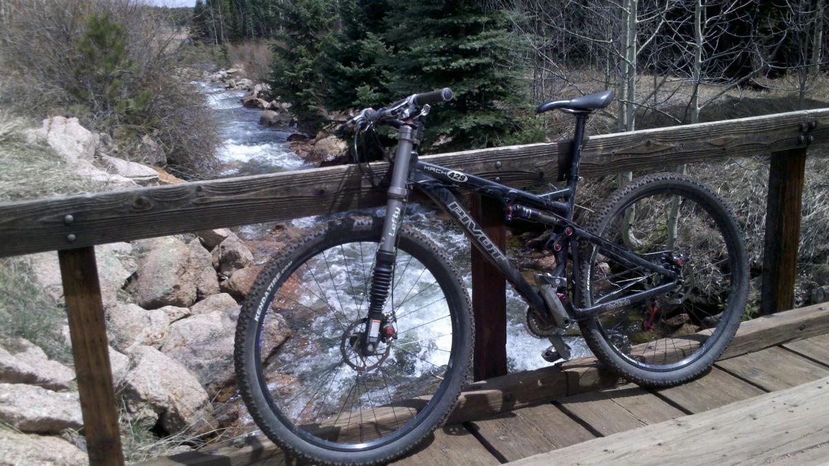 Pivot Mach 429: A mountain bike leaning against a wooden bridge railing with a flowing creek and rocky landscape in the background. Trees and shrubs are visible along the banks, indicating a natural outdoor setting.