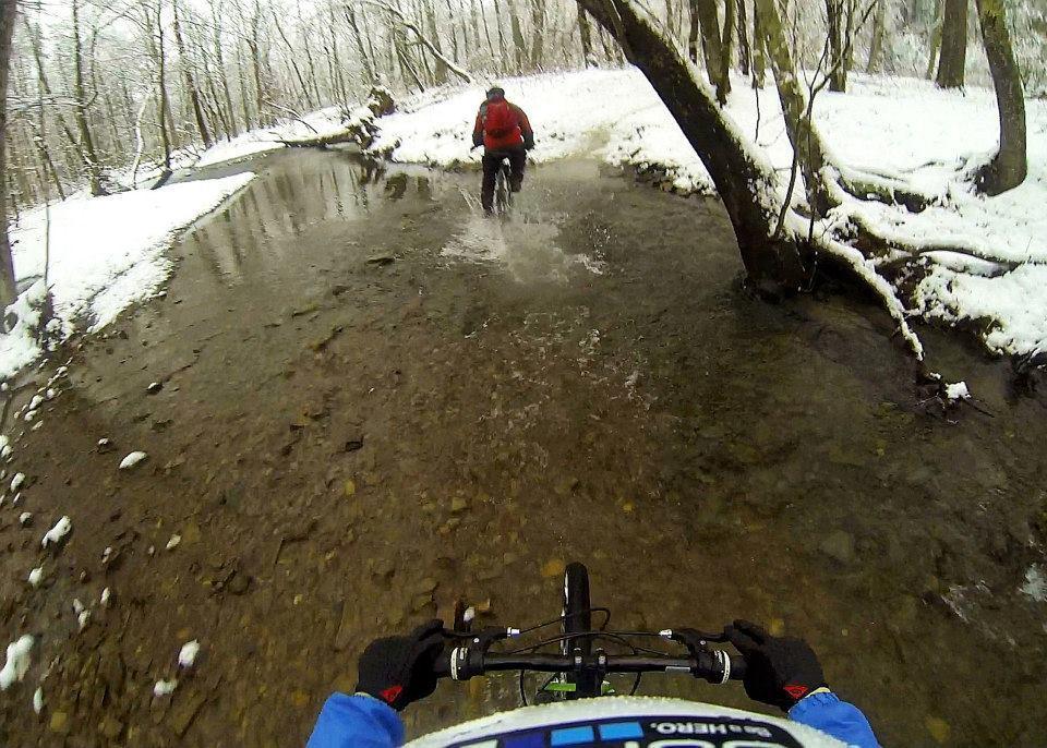A cyclist riding through a shallow stream in a snowy forest. The view is from the cyclist's perspective, showing the bike handlebars and the water splashing around the bike tires. Trees with snow-covered branches line the creek. Comet mountain bike trail.