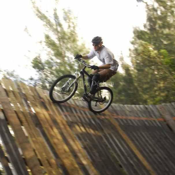A cyclist riding a mountain bike down a wooden ramp in a forested area, showcasing an action shot of the rider in motion. The background features trees and natural light filtering through the leaves. Markham Park mountain bike trail.
