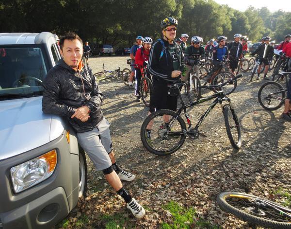 A group of mountain bikers gathered in a parking area, with one individual leaning against a vehicle in the foreground. They are surrounded by trees and gravel, and several riders are preparing their bikes. The scene captures a moment before a biking event or group ride.