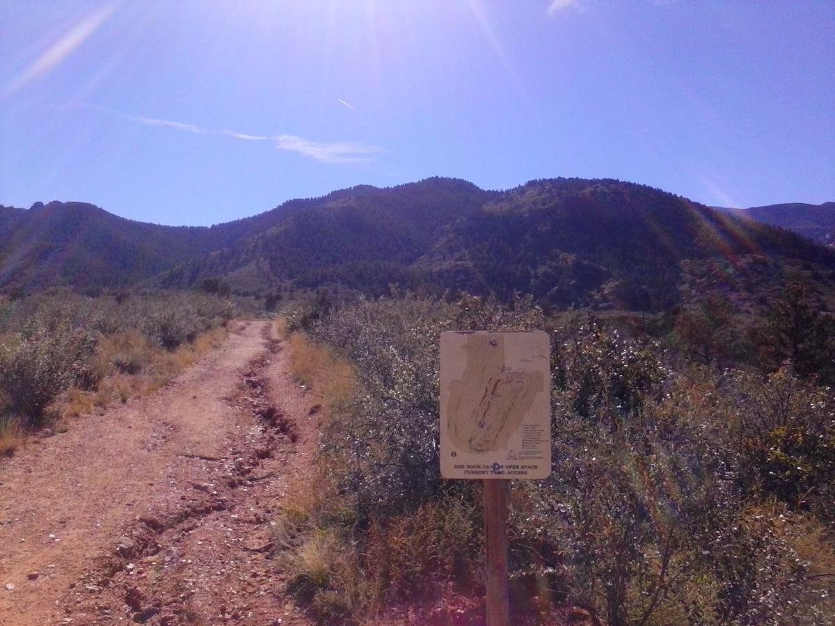 A dirt path leading into a mountainous area, flanked by shrubs and grass. In the foreground, a trail map sign provides information about the area. The sky is clear with bright sunlight shining, and the mountains in the background are partially covered with trees. Red Rock Canyon mountain bike trail.