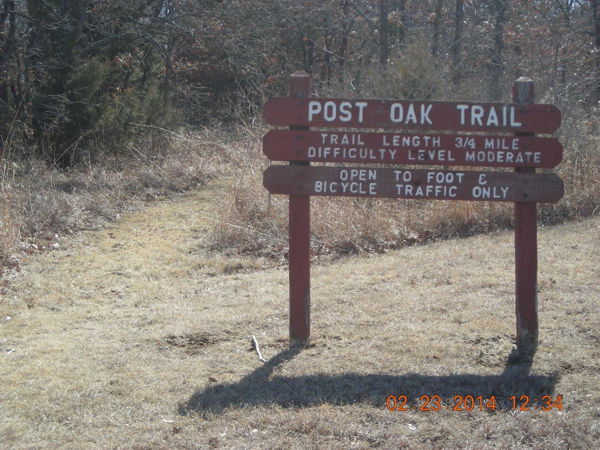 Sign for Post Oak Trail indicating that it is 3/4 mile long, has a moderate difficulty level, and is open for foot and bicycle traffic only. The trail entrance is visible with surrounding grassy areas and sparse trees. Fall River State Park mountain bike trail.