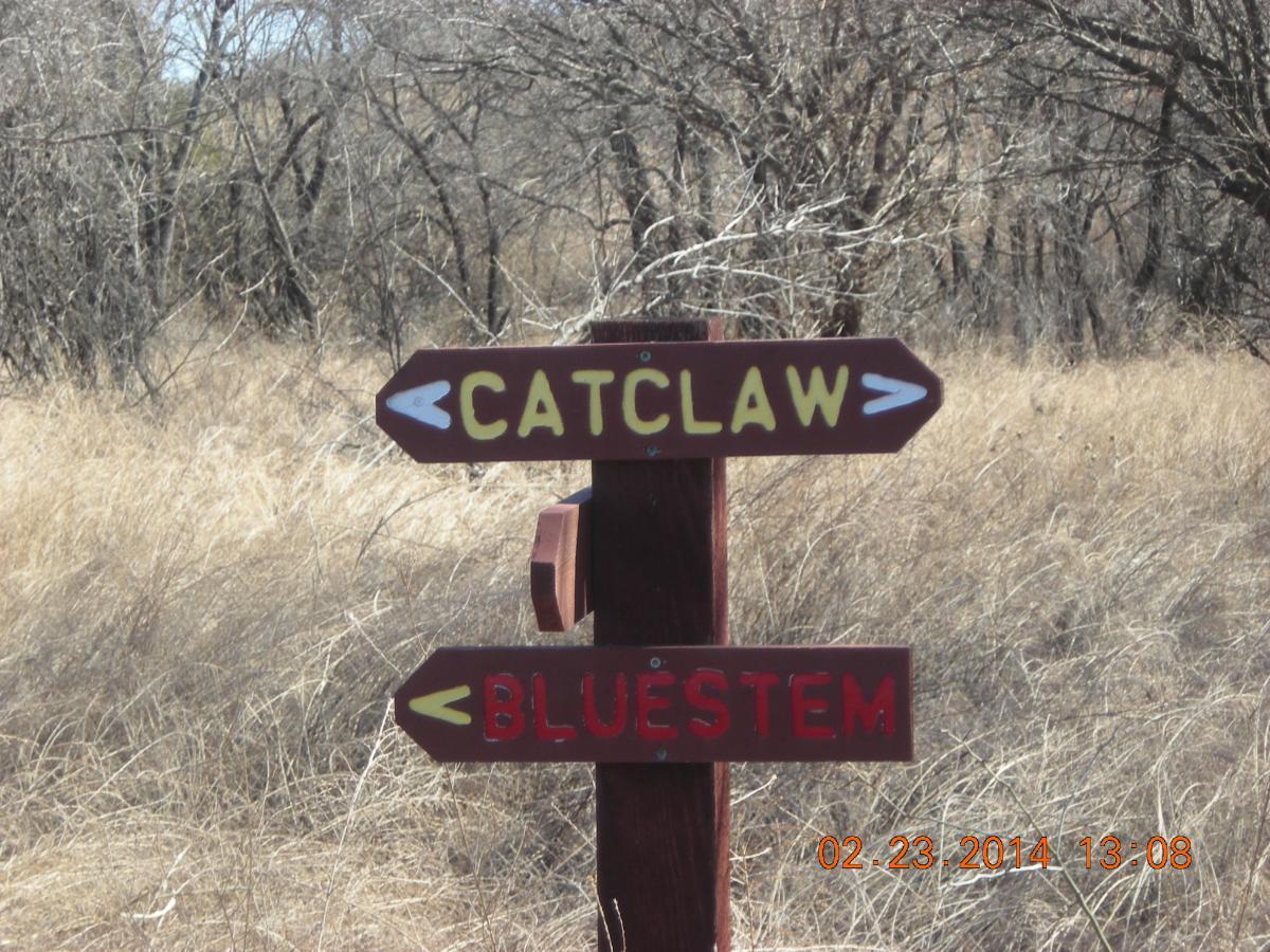 A wooden signpost with two arrows pointing in different directions, labeled "CATCLAW" to the left and "BLUESTEM" to the right, set against a backdrop of dry grass and sparse trees. Fall River State Park mountain bike trail.
