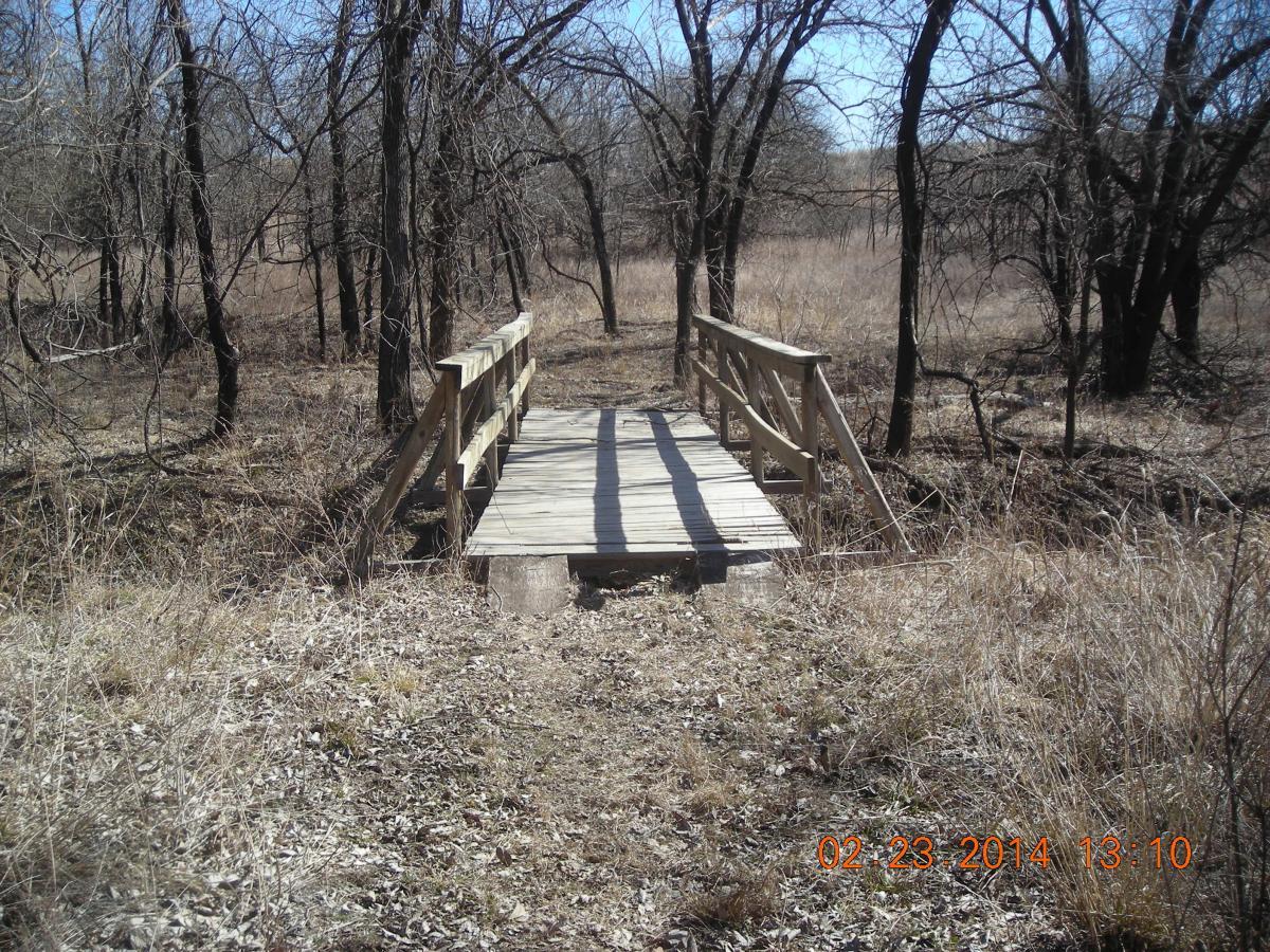 A wooden footbridge crossing over a small area of grass and dried vegetation, surrounded by bare trees in a tranquil outdoor setting. Fall River State Park mountain bike trail.