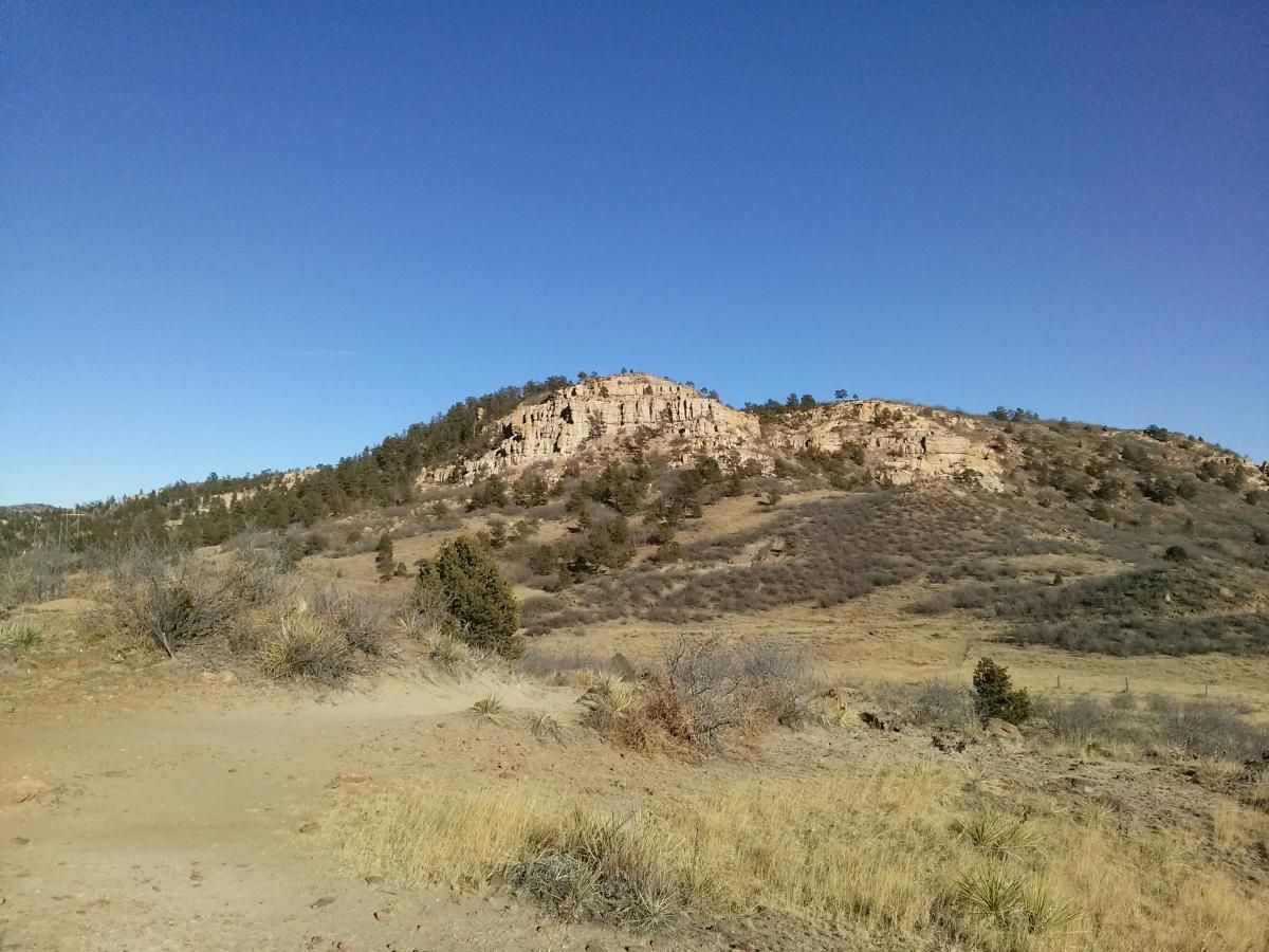A scenic view of a rocky hill surrounded by dry grasses and sparse trees under a clear blue sky. The terrain features gently sloping hills and rocky cliffs in the background. Palmer Park mountain bike trail.