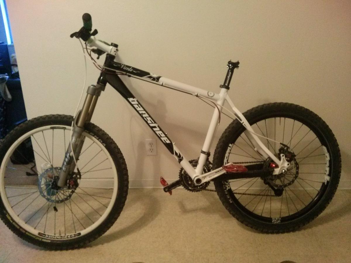 Banshee Viento: A mountain bike with a black and white frame, featuring thick tires for off-road riding. The bike has a front suspension fork, disc brakes, and a multi-gear setup, displayed against a plain wall in an indoor setting.