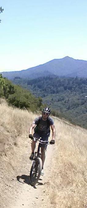 A person riding a mountain bike on a dirt trail surrounded by grassy hills and trees, with a mountain visible in the background under a clear blue sky. Camp Tamarancho mountain bike trail.