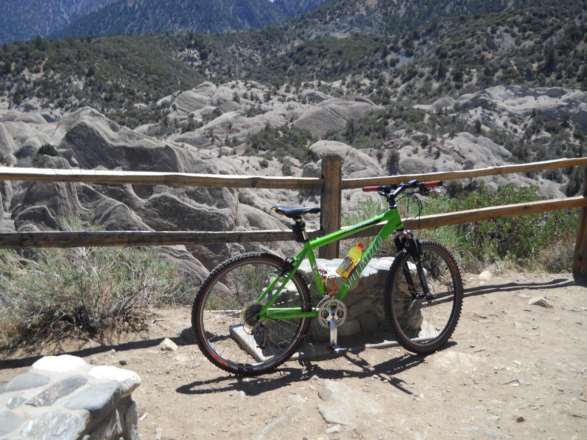 Specialized Rockhopper: A bright green mountain bike is parked near a rocky landscape, surrounded by shrubs and a wooden fence. The background features rugged, mountainous terrain under a clear blue sky.