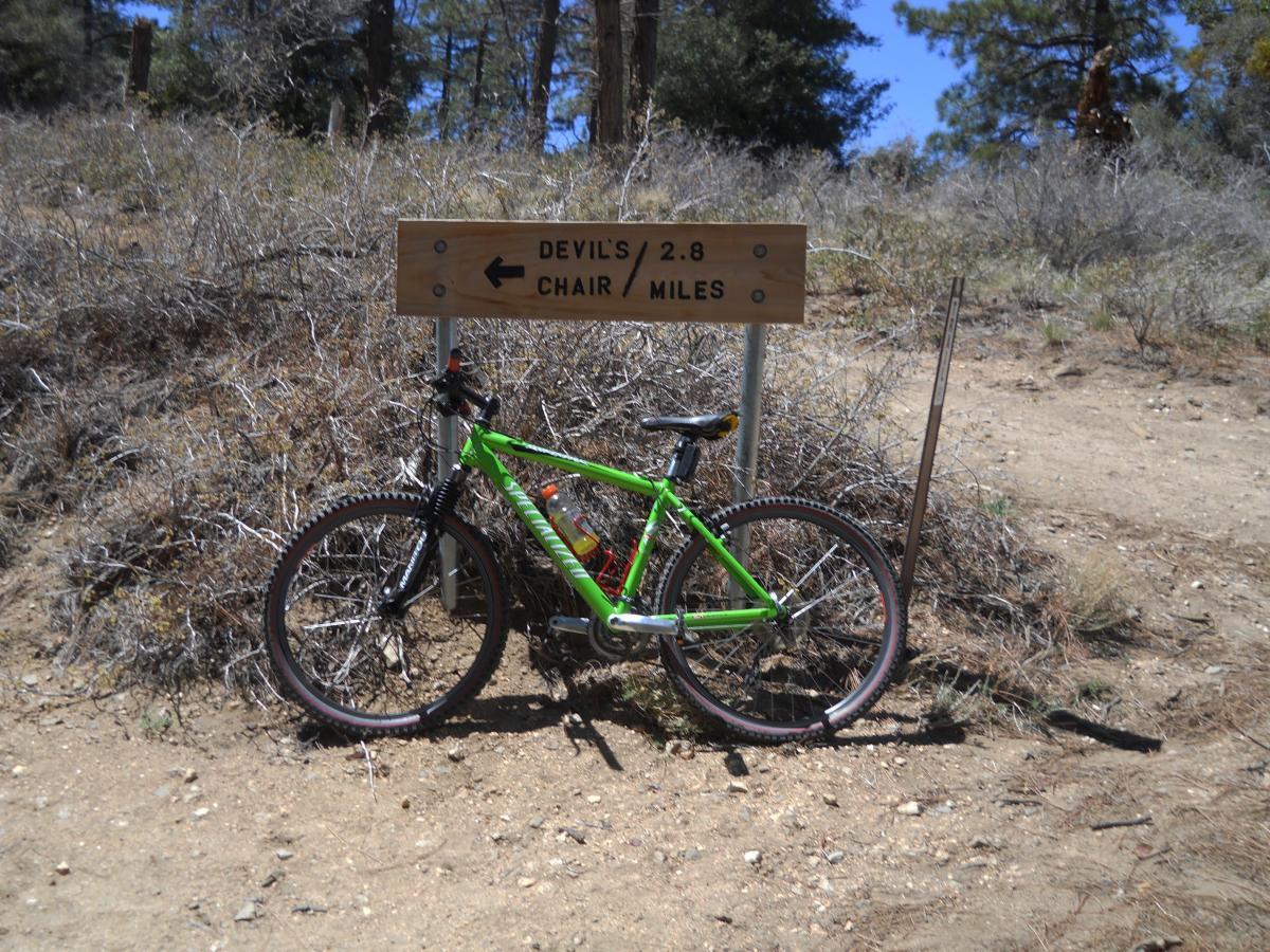 Specialized Rockhopper 29: A bright green mountain bike parked next to a wooden trail sign indicating directions to "Devil's Chair" and "2.8 Miles." Surrounding the bike is a dry, natural landscape with sparse vegetation and dirt pathways.