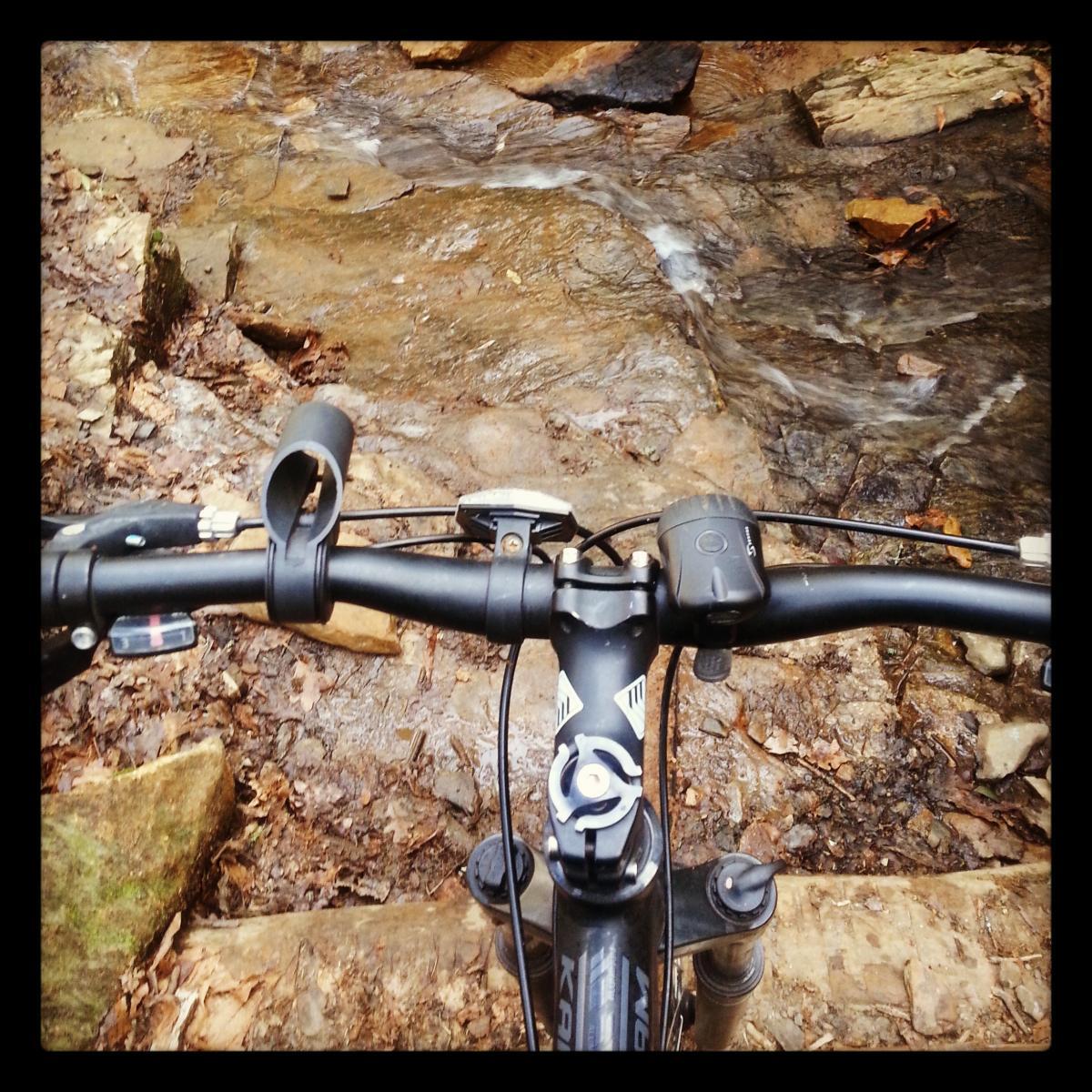 A close-up view of a mountain bike's handlebars, positioned above a rocky path with a small stream flowing nearby. The ground is covered with leaves and stones, showcasing a natural outdoor setting ideal for biking. Reasonover Creek Trail mountain bike trail.