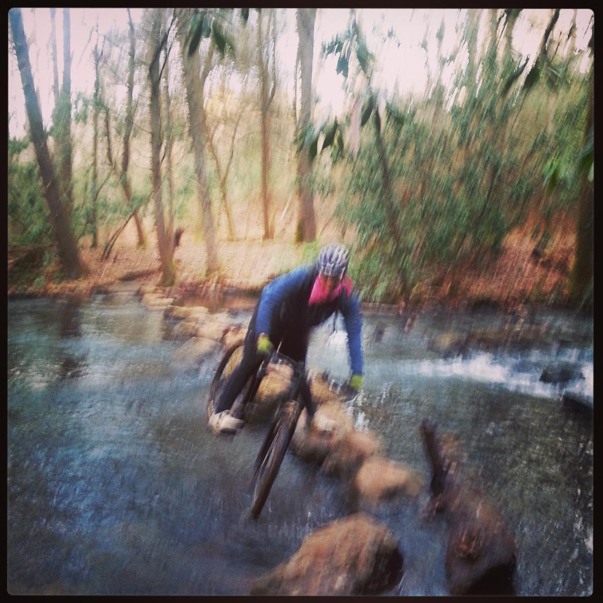 A cyclist navigating rocky terrain while crossing a stream in a wooded area, with blurred motion conveying speed and excitement. Reasonover Creek Trail mountain bike trail.