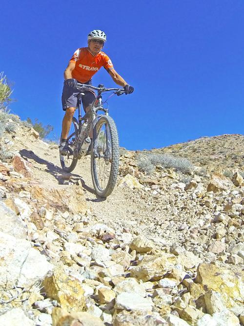 A person riding a mountain bike on a rocky trail surrounded by desert terrain. The cyclist is wearing an orange shirt and a helmet, navigating a steep incline under a clear blue sky. Bootleg Canyon mountain bike trail.