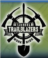 Logo for Tarheel Trailblazers featuring a circular design with a bicycle wheel in the center, surrounded by crossed tools including a pickaxe, shovel, and a hammer. The words "TARHEEL TRAILBLAZERS" are prominently displayed, along with the motto "WORK RIDE." The overall color scheme includes shades of green and black.