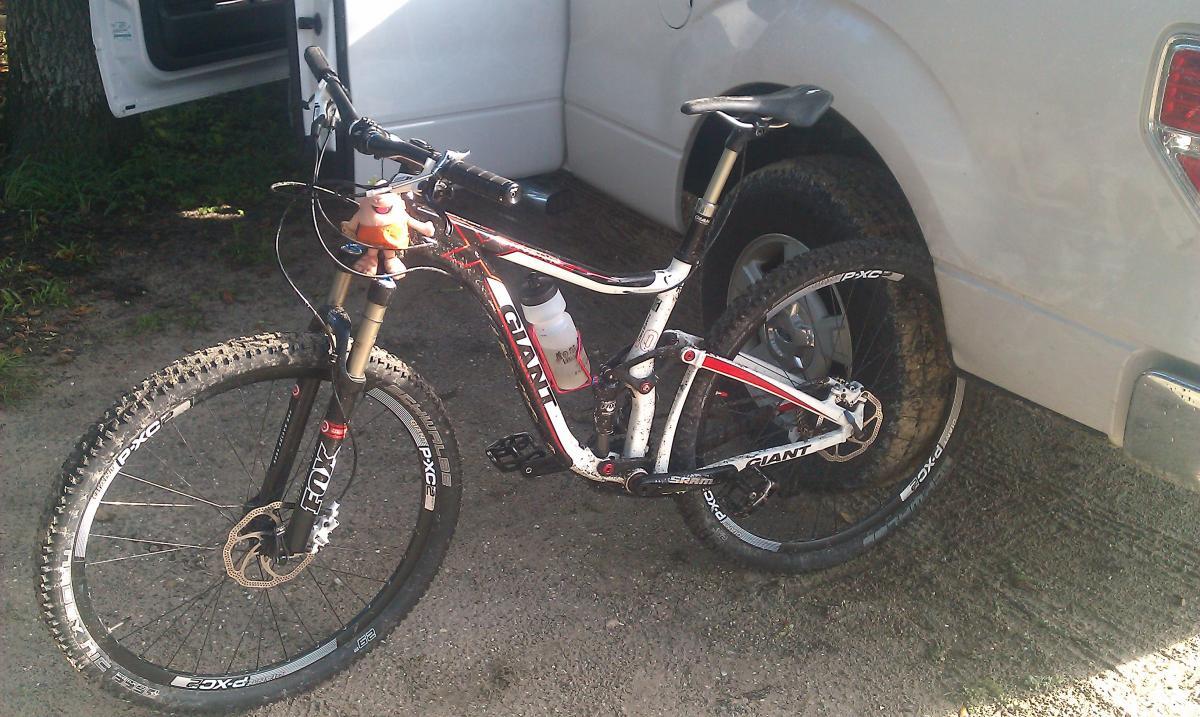 Giant Trance X 29er 1: A black and white mountain bike with red accents parked next to a white vehicle. The bike has a water bottle attached and features knobby tires, indicating it is designed for off-road riding. The background shows a dirt surface and greenery, suggesting an outdoor setting.