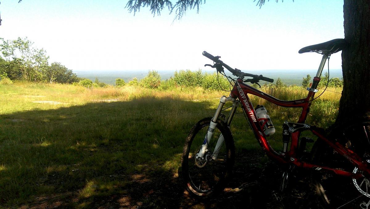 Rocky Mountain Slayer 50: Mountain bike resting in the foreground with a scenic view of lush green fields and a distant horizon under a clear sky.