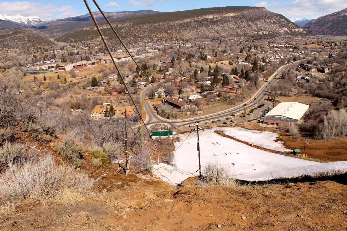 A panoramic view from the top of a hill overlooking a small town surrounded by mountains. In the foreground, there is a ski area with patches of snow and ski lifts. The town features various buildings, roads, and trees, with a backdrop of mountains in the distance under a partly cloudy sky. Horse Gulch mountain bike trail.