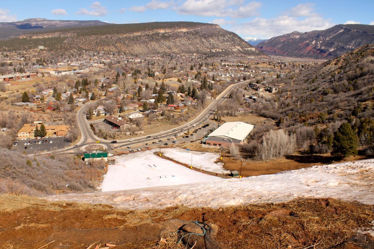 A scenic view overlooking a small town nestled in a valley, surrounded by mountains. In the foreground, there is a snow-covered area, while the town features a mix of residential homes and commercial buildings along winding roads. Sparse trees dot the landscape, and the sky is partly cloudy, creating a picturesque backdrop. Horse Gulch mountain bike trail.
