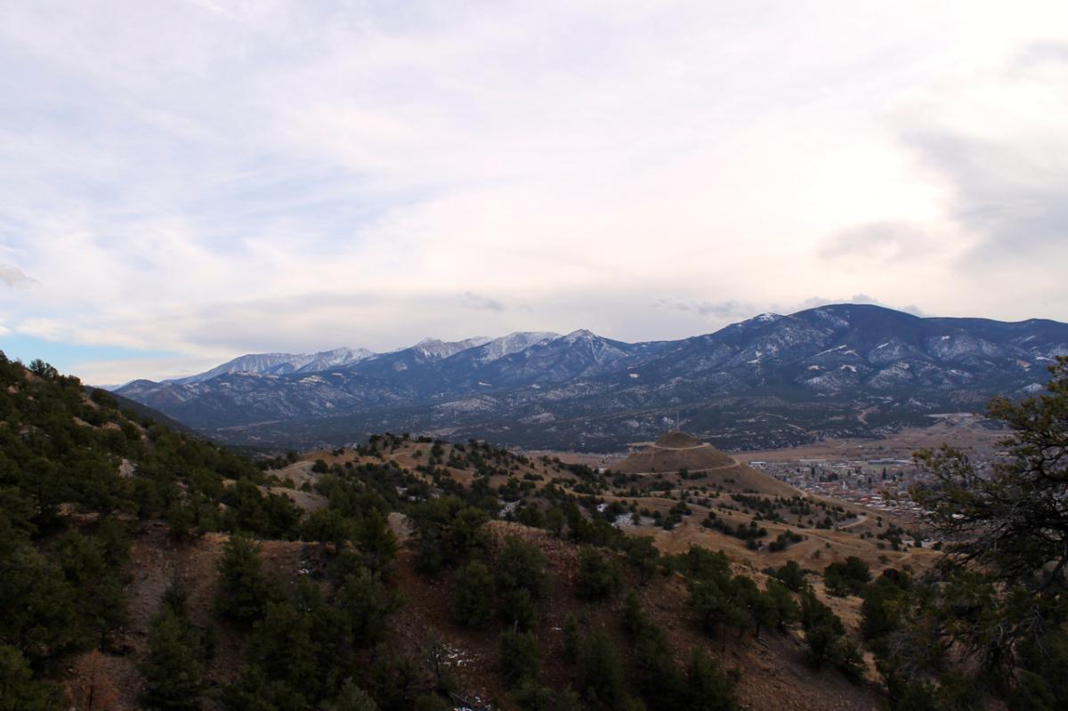 A wide view of a mountainous landscape featuring snow-capped peaks in the background. In the foreground, rolling hills are covered with green pine trees, while a small town is visible in the valley below. The sky is partly cloudy, creating a serene and picturesque atmosphere. North Backbone mountain bike trail.