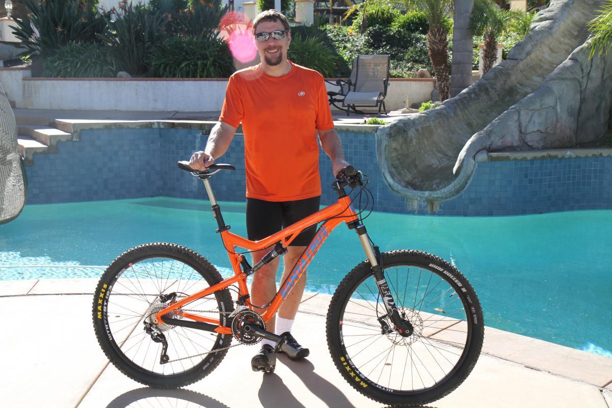 Santa Cruz 5010: A man in an orange shirt and black shorts stands next to a bright orange mountain bike near a pool. The background features lush greenery and a water slide. The sun is shining, creating a bright and vibrant atmosphere.