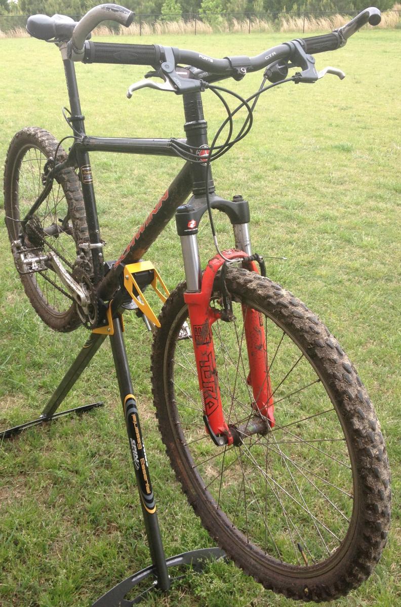 Schwinn Homegrown: Mountain bike displayed on a repair stand, with muddy tires and a black frame featuring red fork components. The background shows a grassy area, indicating an outdoor setting.