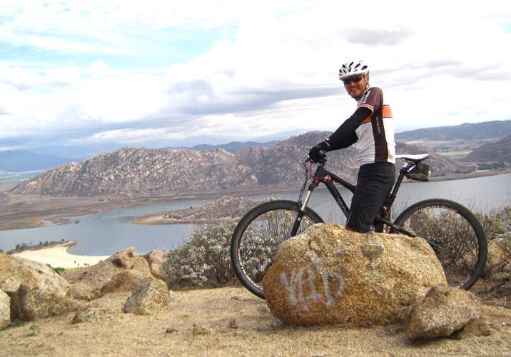 A mountain biker stands on a large rock at a scenic overlook, smiling at the camera. The background features a lake surrounded by hills and mountains under a partly cloudy sky. The biker is wearing a helmet and cycling gear, and the bicycle is positioned next to the rock. Terri Peak mountain bike trail.
