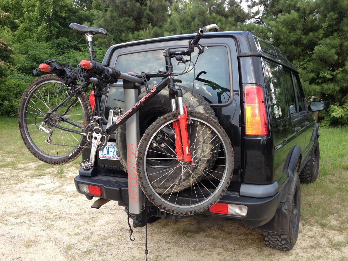 Schwinn Homegrown: Image of a black SUV parked on a gravel surface with a mountain bike mounted on a bike rack attached to the rear. The bike features thick tires and a red accent, and the vehicle shows signs of outdoor activity, surrounded by greenery.