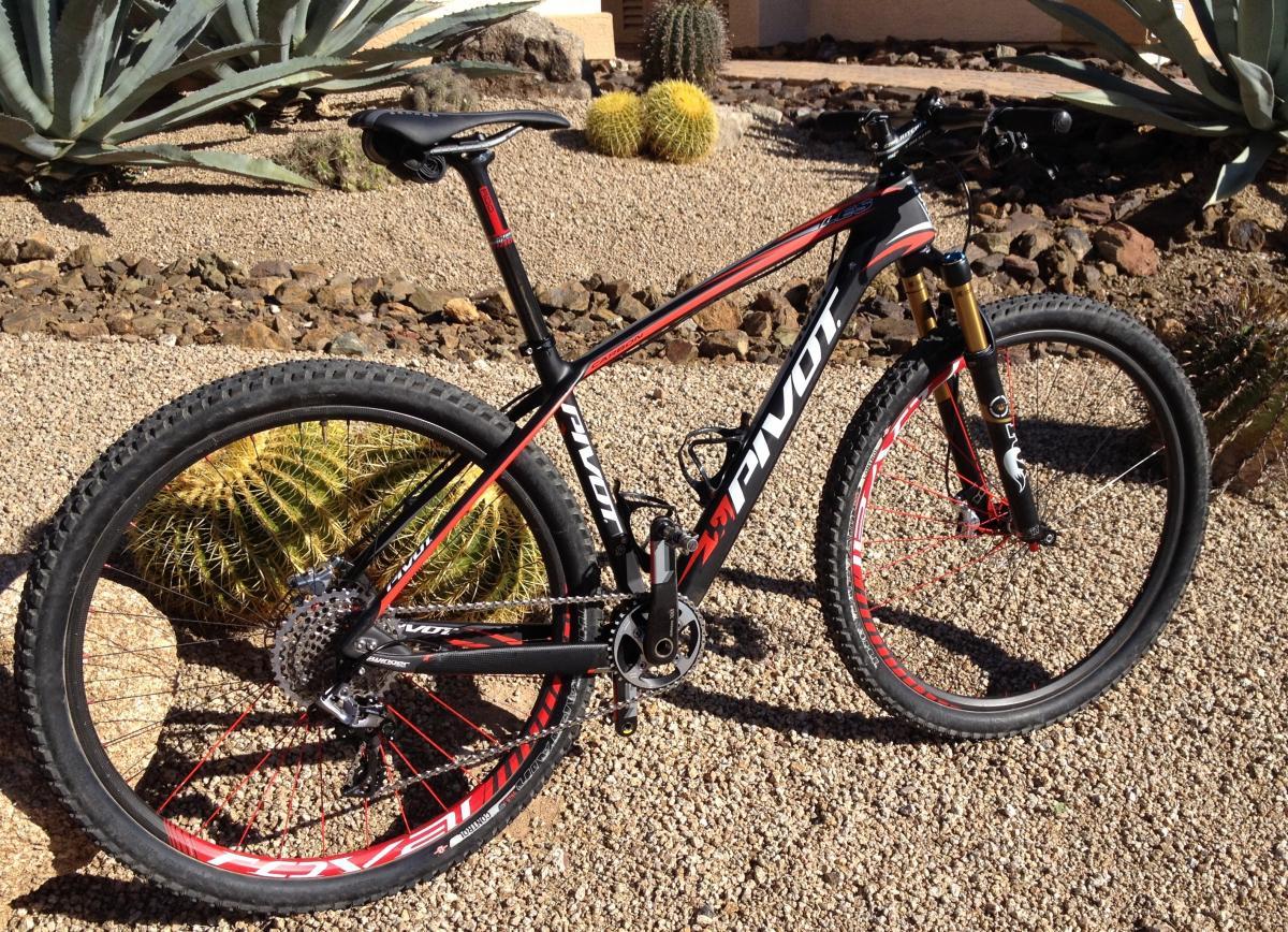 Pivot Les: A black and red mountain bike is parked on a gravel surface, surrounded by desert plants, including cacti and agave. The bike features thick tires, a prominent suspension fork, and a sleek frame design with branding visible.