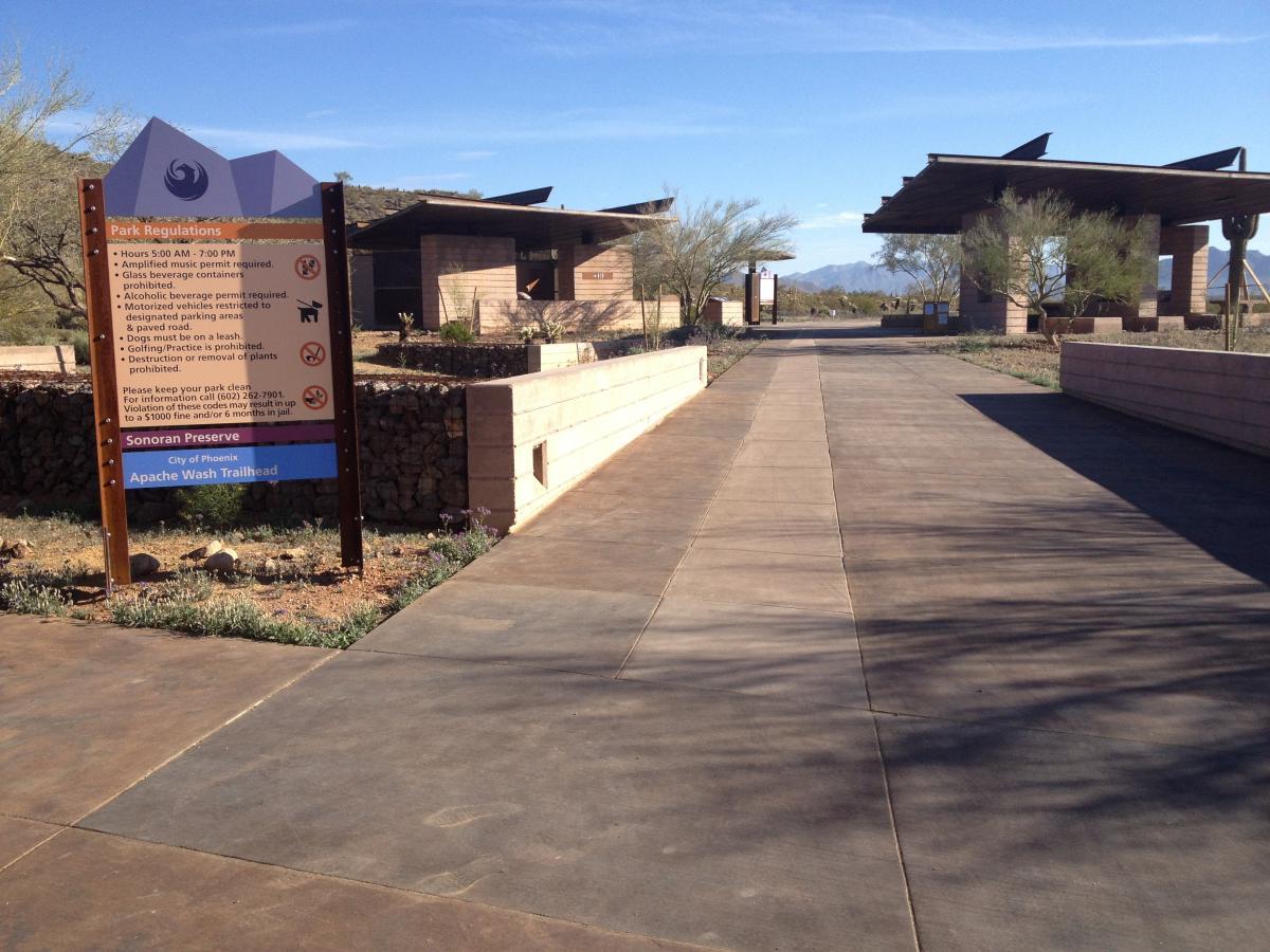 A pathway leading to the Apache Wash Trailhead, featuring a park regulations sign with rules and hours of operation. The landscape includes desert vegetation and modern architectural structures under a clear blue sky. Sonoran Preserve North mountain bike trail.