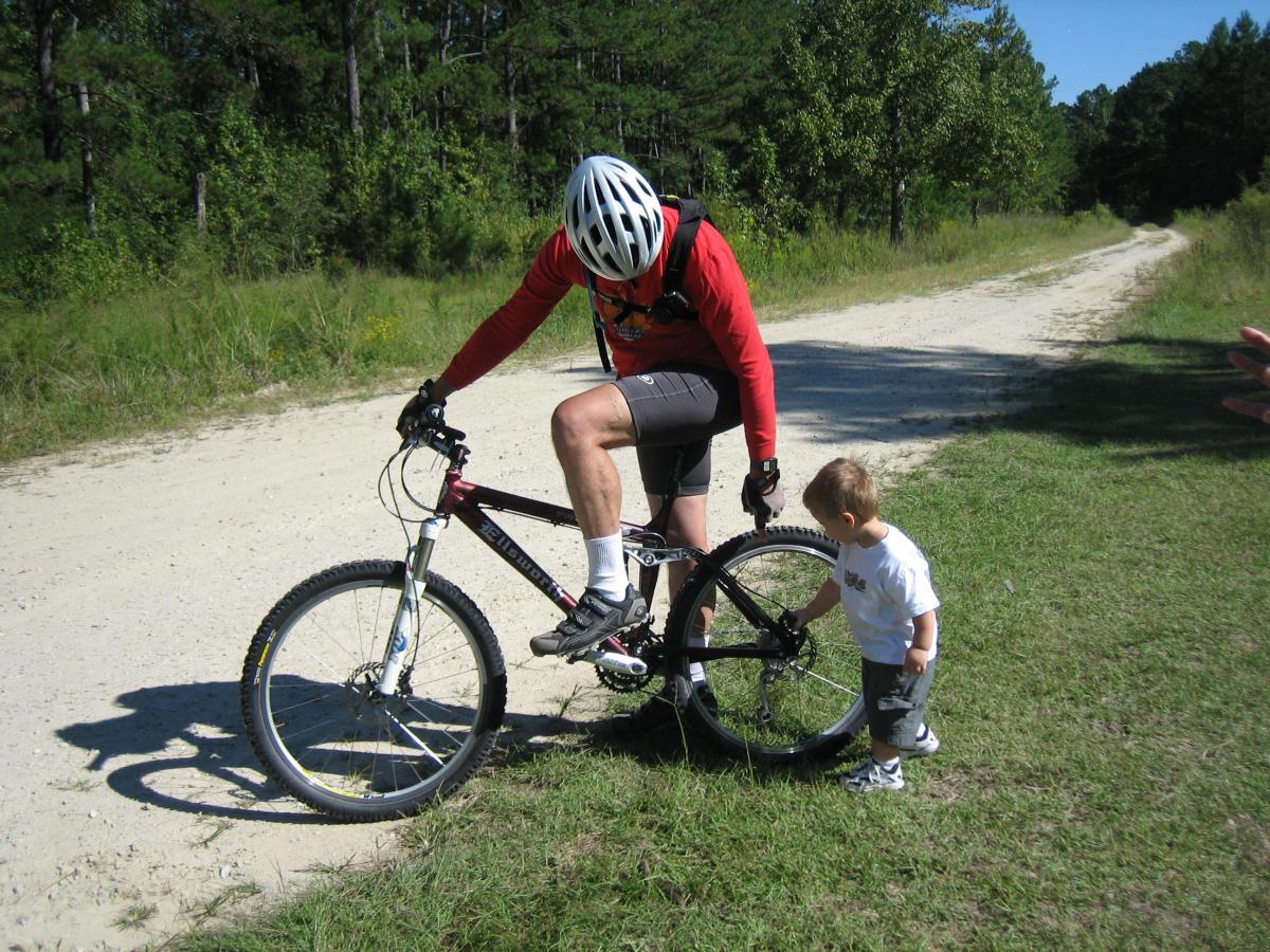 Ellsworth Truth: A person in a red long-sleeve shirt and a bike helmet is crouching beside a mountain bike on a dirt path, examining the bike. A young child in a white shirt and gray shorts is standing nearby, reaching out toward the bike. Lush greenery and trees are visible in the background, indicating a wooded area.