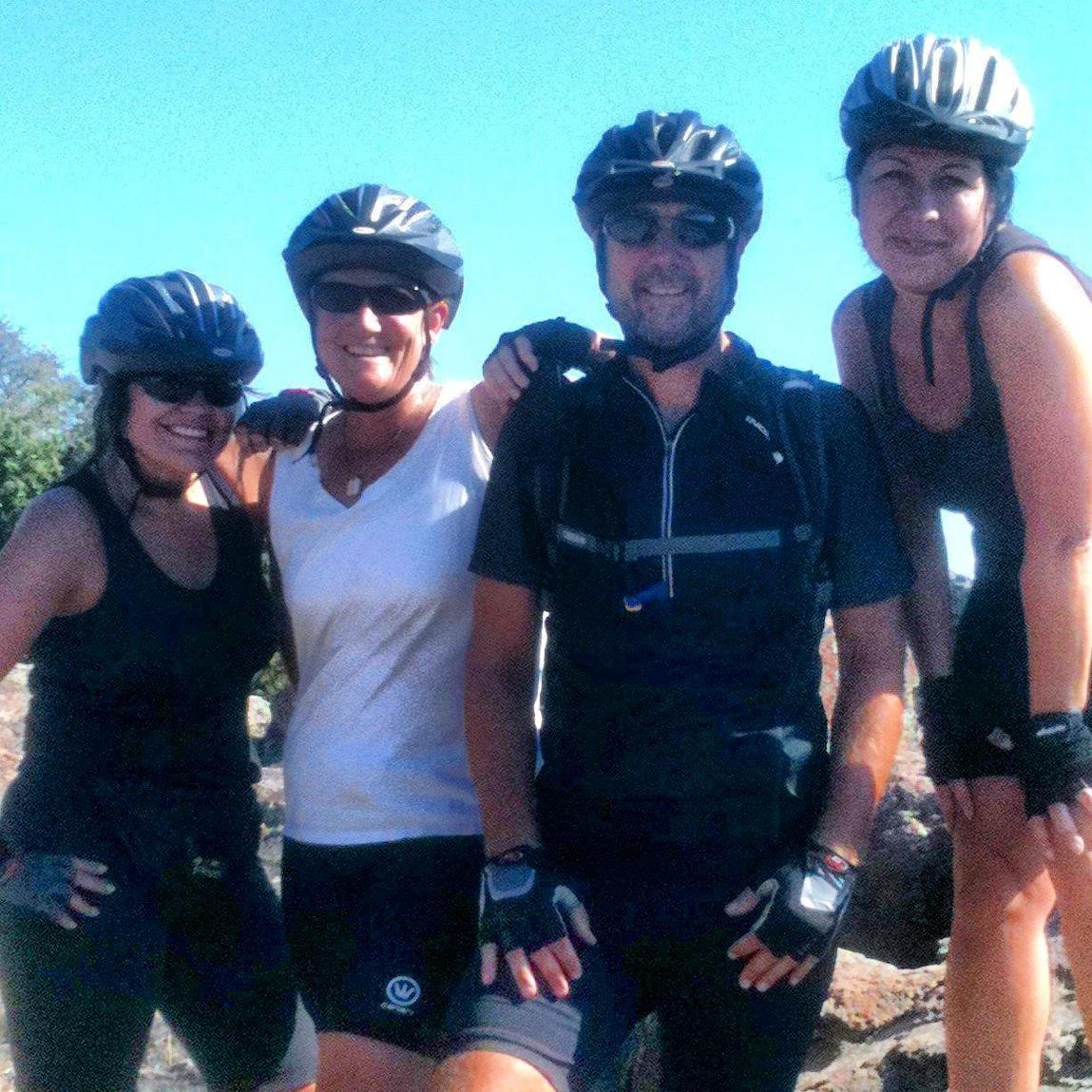 Four cyclists wearing helmets and athletic clothing smile for the camera against a clear blue sky. They appear to be enjoying a break during a bike ride, standing in a rocky outdoor setting. Rockville Park mountain bike trail.