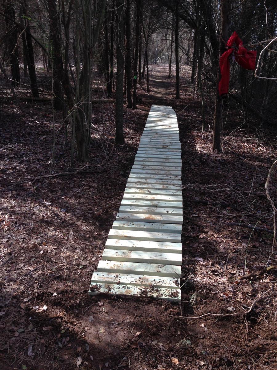 A wooden boardwalk stretches through a wooded area, lined with trees and leaf litter on the ground. The boardwalk is made of planks, providing a pathway through the natural environment. A red piece of cloth is visible hanging from a tree branch in the background. Spadra Creek Nature Trail mountain bike trail.
