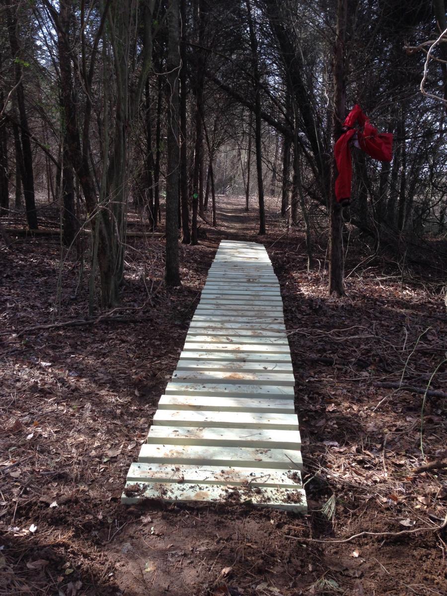 A wooden path stretching through a forest, surrounded by tall trees and dry leaves on the ground. A red piece of clothing is hung on a tree branch to the right of the path. Spadra Creek Nature Trail mountain bike trail.