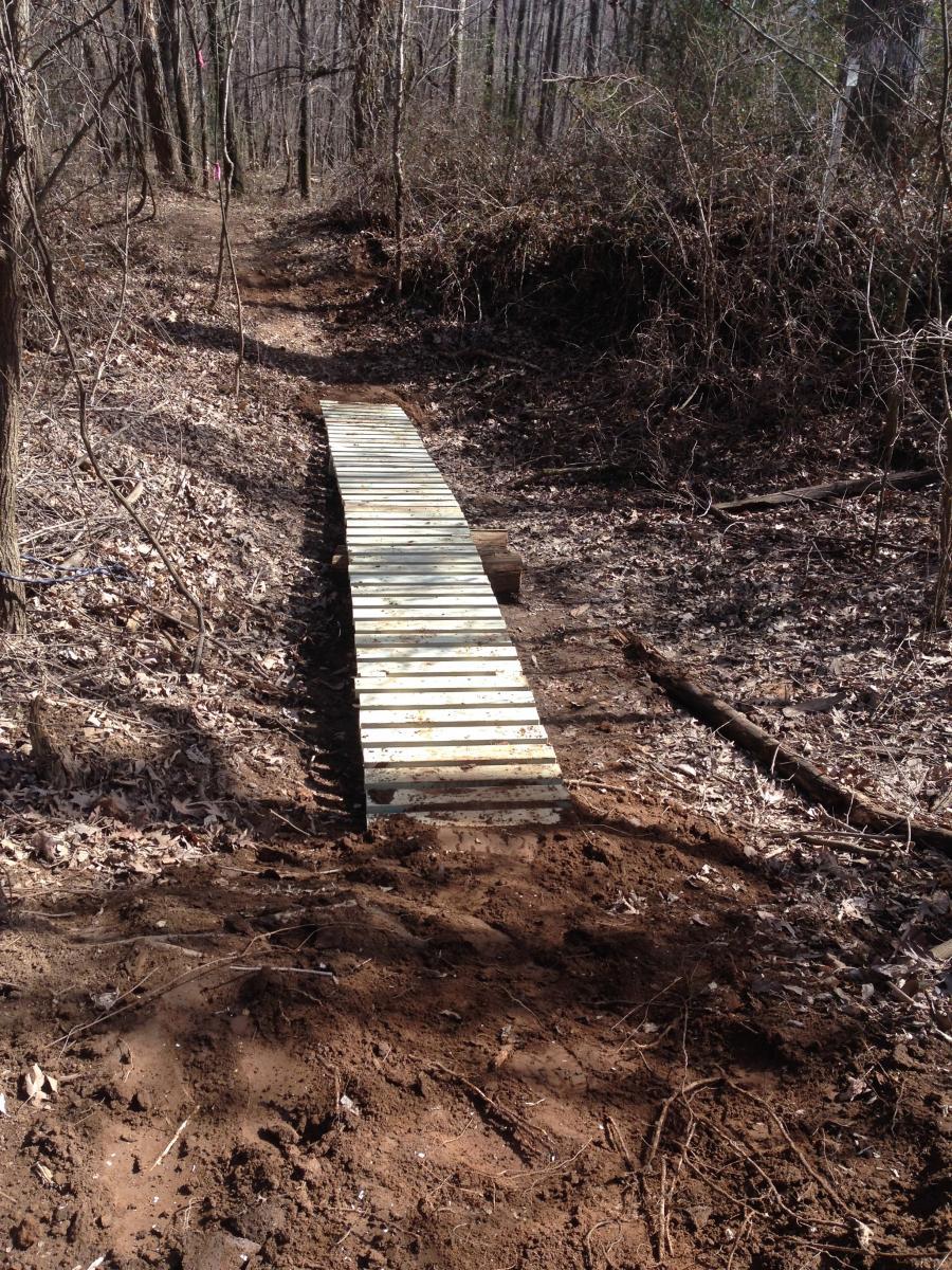 A wooden bridge spans a narrow trail in a wooded area, surrounded by leafless trees and underbrush. The ground is covered with dry leaves and exposed earth, indicating a natural, unpaved hiking path. Spadra Creek Nature Trail mountain bike trail.