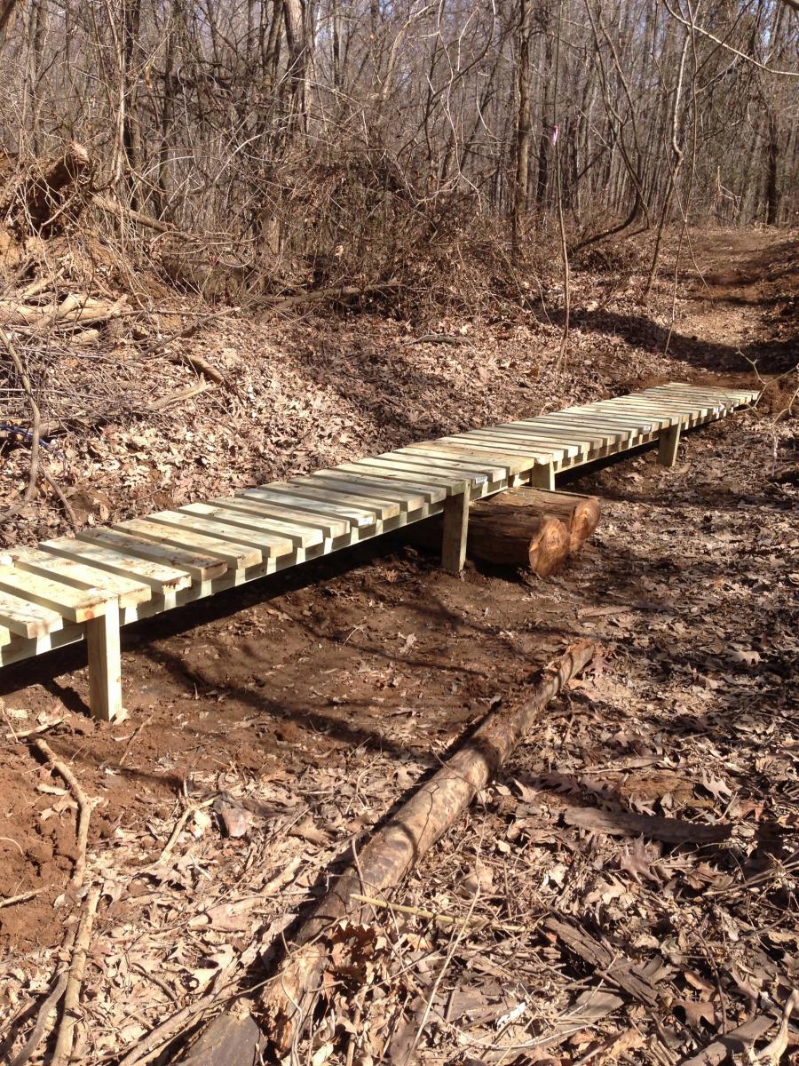 A wooden walkway extends over a muddy path in a wooded area, surrounded by bare trees and dry leaves. The structure is made of planks and is supported by logs, providing a stable passage through the natural landscape. Spadra Creek Nature Trail mountain bike trail.