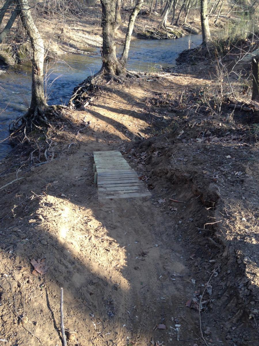 A narrow dirt path leading to a small wooden bridge over a stream, surrounded by trees. The area is slightly uneven with exposed roots and leaf litter, indicating a natural setting. Sunlight casts shadows on the ground, highlighting the texture of the soil and vegetation. Spadra Creek Nature Trail mountain bike trail.