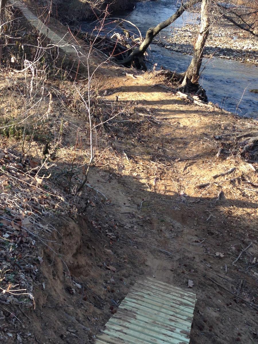 A dirt path leading down to a stream, with a wooden bridge crossing a section of the path in the foreground. The surrounding area features sparse vegetation and bare trees, indicating a late autumn or early winter setting. Spadra Creek Nature Trail mountain bike trail.