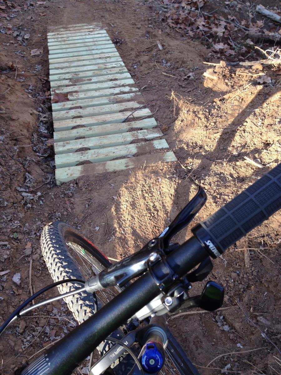 A close-up view from the handlebars of a mountain bike, showing a narrow wooden bridge made of planks on a dirt trail. The surrounding area is earthy with some scattered leaves and dirt, indicating an outdoor setting ideal for biking. Spadra Creek Nature Trail mountain bike trail.