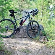 KHS Winslow: A mountain bike with a helmet placed on it, resting on a rocky area surrounded by greenery and trees.