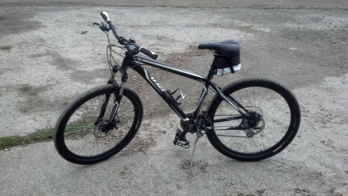 Specialized Hardrock Disc: A black mountain bike parked on a gravel surface, with thick tires and a small saddle bag attached to the seat. The surrounding area appears slightly overgrown with grass.