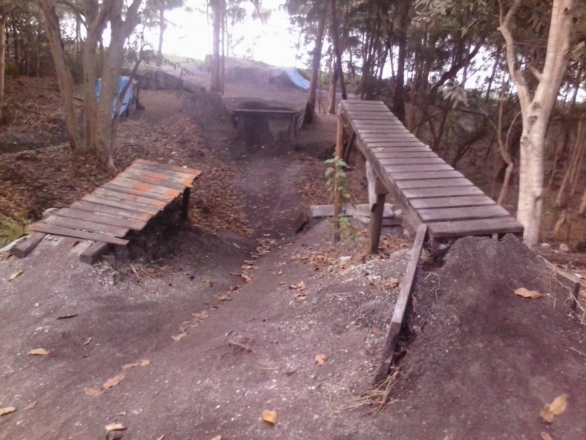 A dirt bike area featuring two wooden ramps positioned across a dirt track in a wooded environment. The ramps are made of wooden planks and are situated on either side of a small mound, with fallen leaves scattered on the ground. The background includes trees and hints of blue tarps in the distance. Markham Park mountain bike trail.