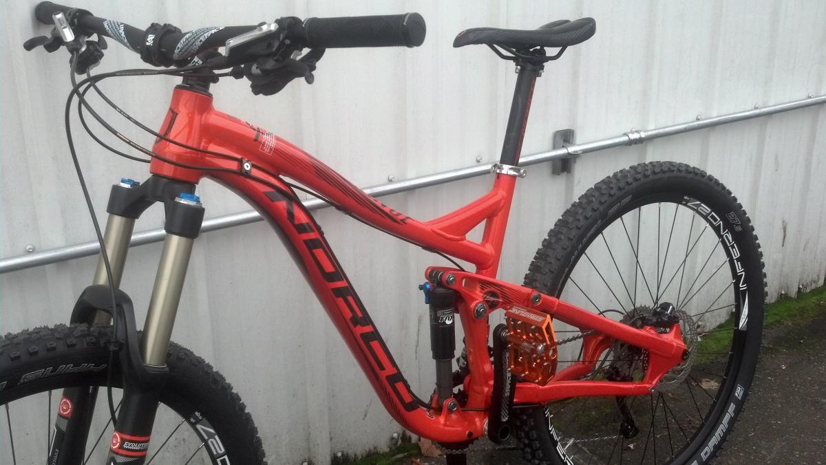 Norco Range 7.1: A close-up view of a vibrant orange mountain bike with a sturdy aluminum frame, featuring a dual suspension system, front forks, and thick tires designed for rugged terrain. The bike is positioned against a textured white wall, with visible brake and gear components showcasing its performance features.
