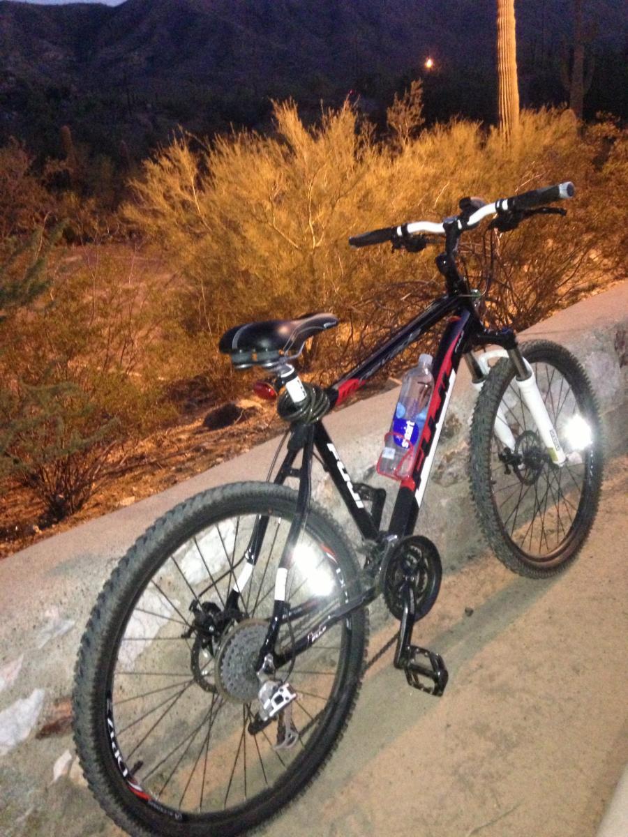 Fuji Nevada 1.0: A black mountain bike parked on a stone wall, with a water bottle attached to the frame. The background features desert vegetation and distant mountains at dusk, with soft lighting illuminating the scene.