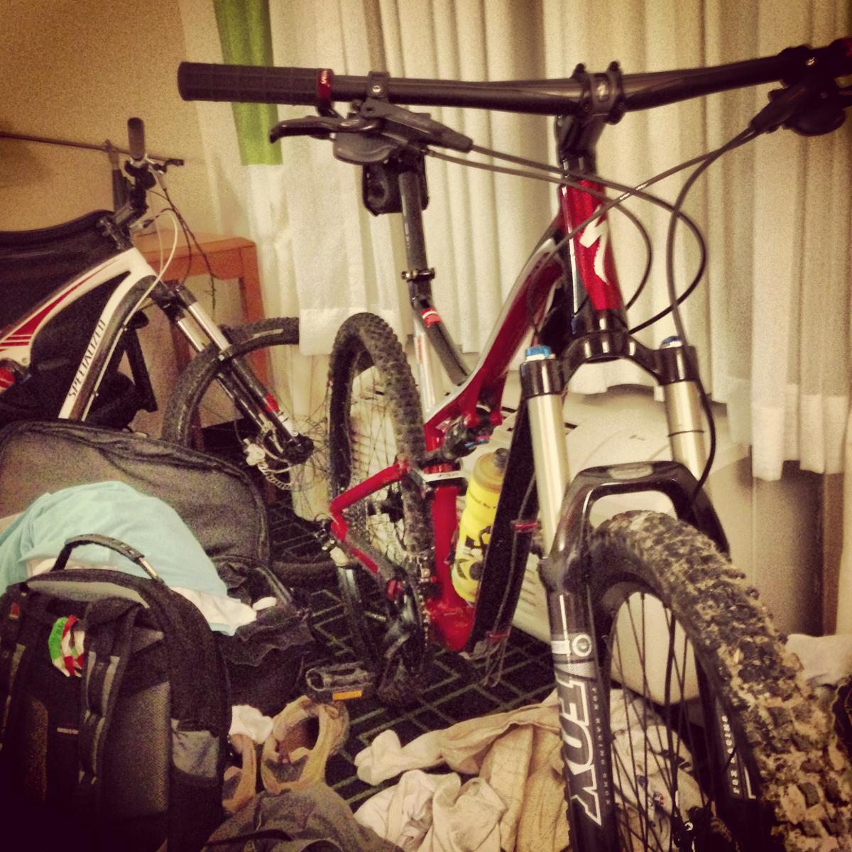 Specialized Stumpjumper FSR Comp 29: A cluttered hotel room featuring two mountain bikes, one red and one white, among scattered clothing and bags on the floor. The bikes show signs of recent use, with dirt on their tires. Vertical blinds are partially drawn in the background.