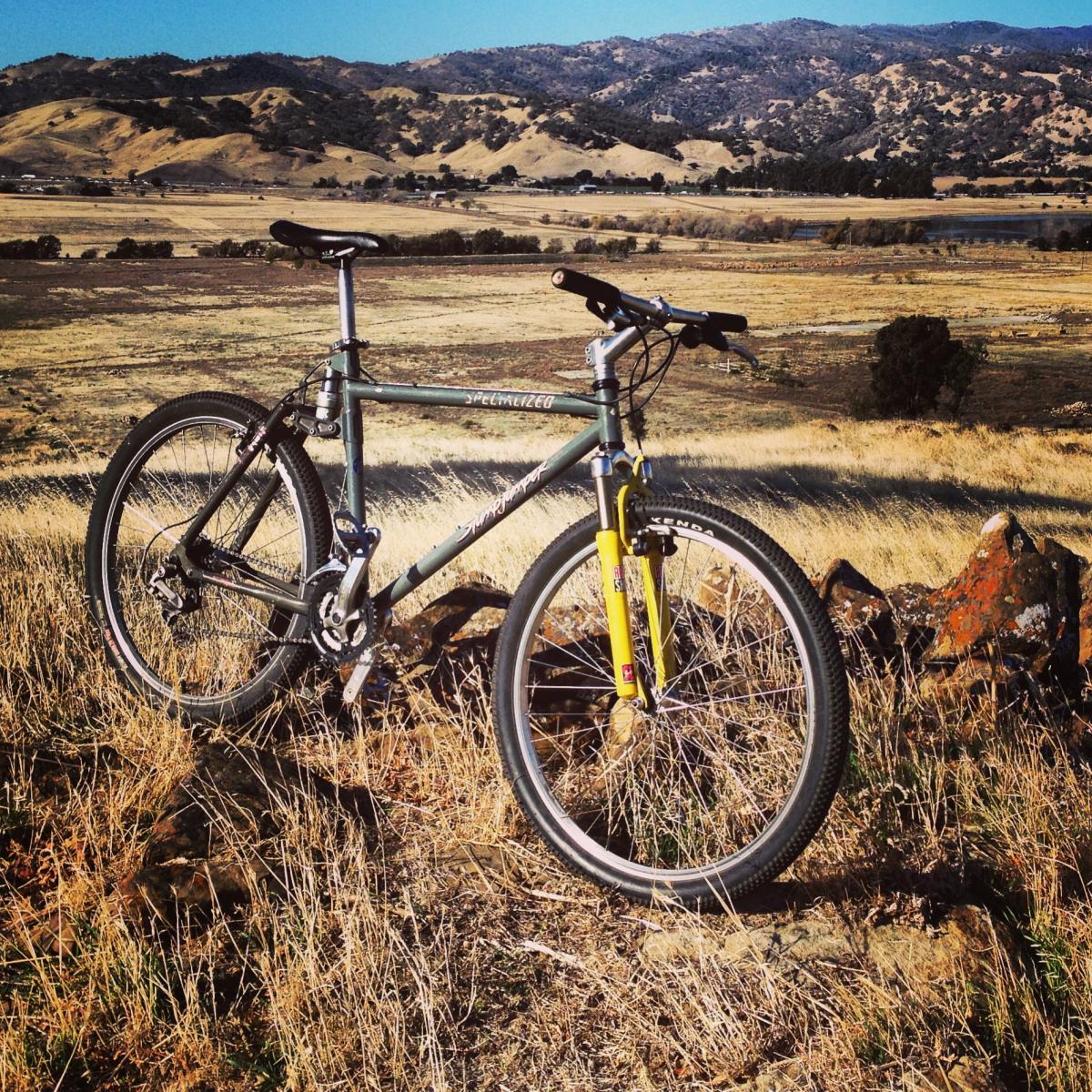 Specialized Stumpjumper: A mountain bike parked on a rocky outcrop, with rolling hills in the background and dry grass in the foreground. The bike has a green frame with yellow front suspension and is set against a clear blue sky.