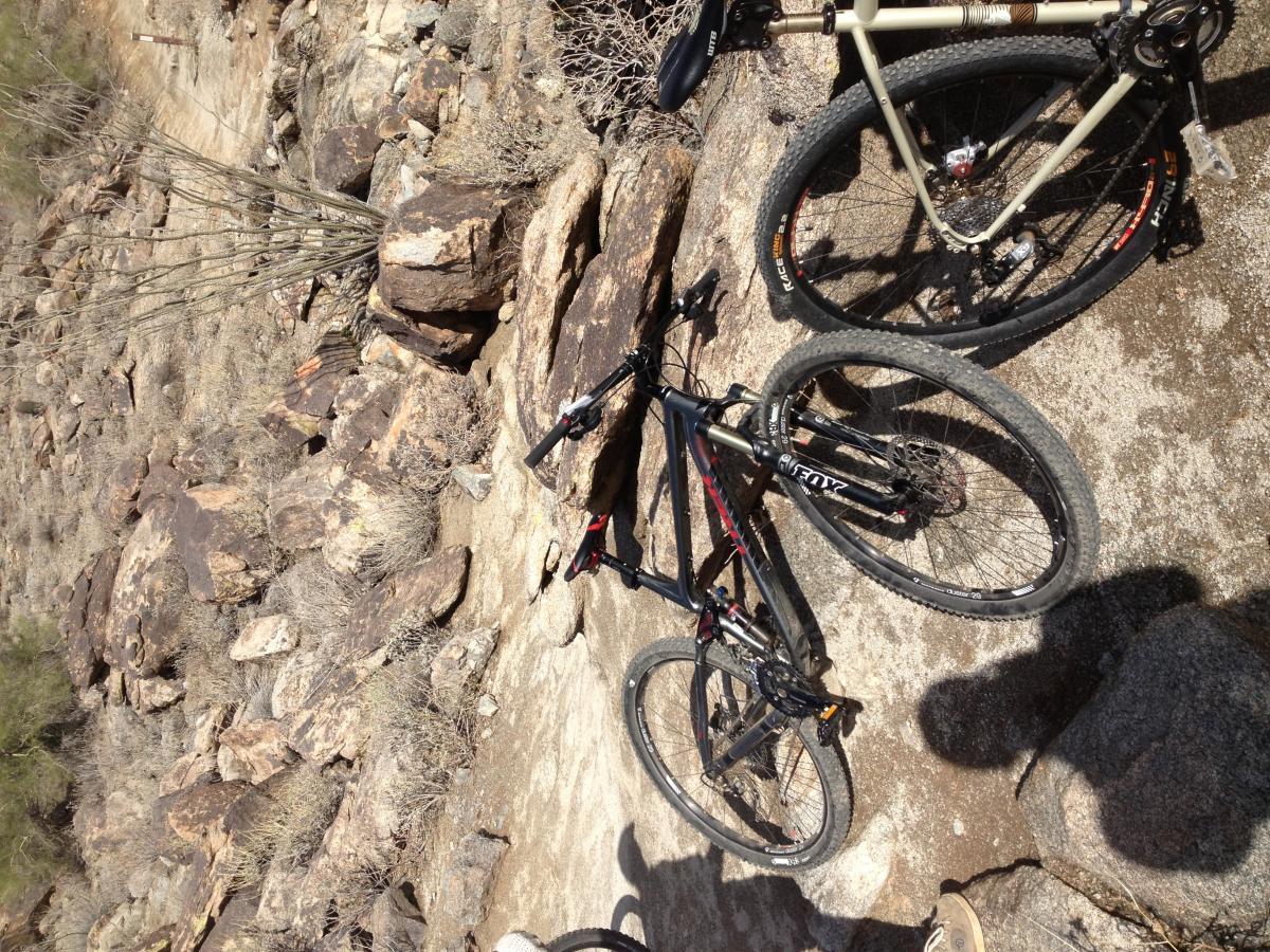 Trek Fuel EX 8 29: Two mountain bikes are positioned on rocky terrain surrounded by sparse vegetation. The bike on the left is black with red accents, while the bike on the right is a lighter color. The background features a mix of large rocks and dry brush, indicative of a rugged outdoor setting.