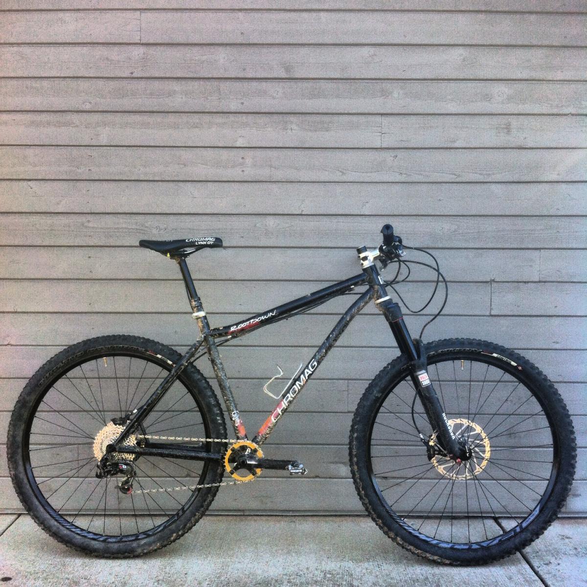 Chromag Rootdown: A black mountain bike with a sturdy frame and thick tires is leaning against a gray wooden wall. The bike features a simplistic design, with visible components such as the handlebars, seat, and gears, showcasing signs of use and dirt.