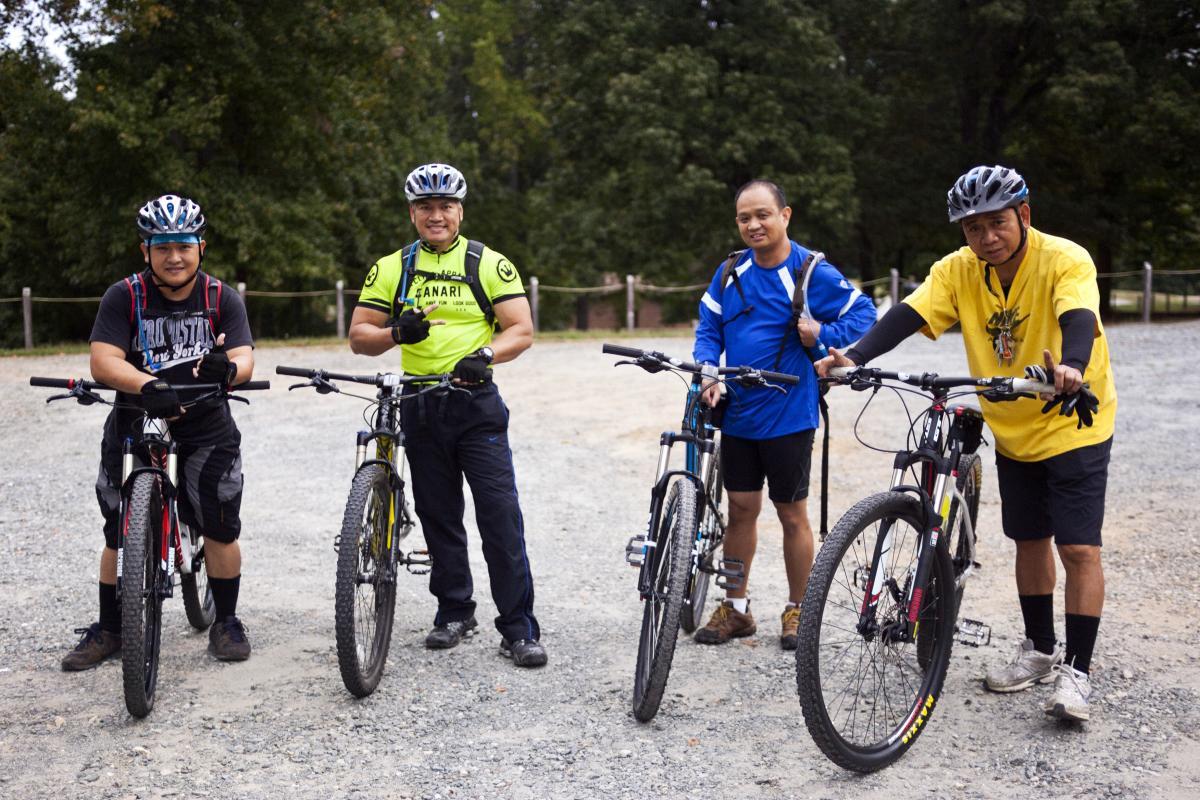 Four men standing with mountain bikes on a gravel path surrounded by trees. They are smiling and wearing cycling gear, including helmets. One man is dressed in a bright yellow shirt, two are in blue tops, and one is wearing a black t-shirt. Hobby Park mountain bike trail.