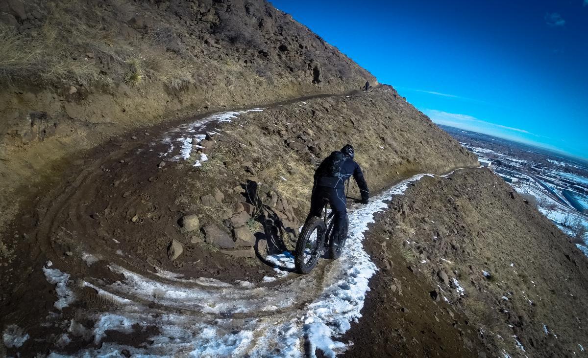 A person riding a fat bike along a narrow, winding dirt trail on a hillside, with patches of snow visible on the ground, under a clear blue sky. The landscape features rocky terrain and sparse vegetation, with a view of a valley in the background. North Table Mountain mountain bike trail.